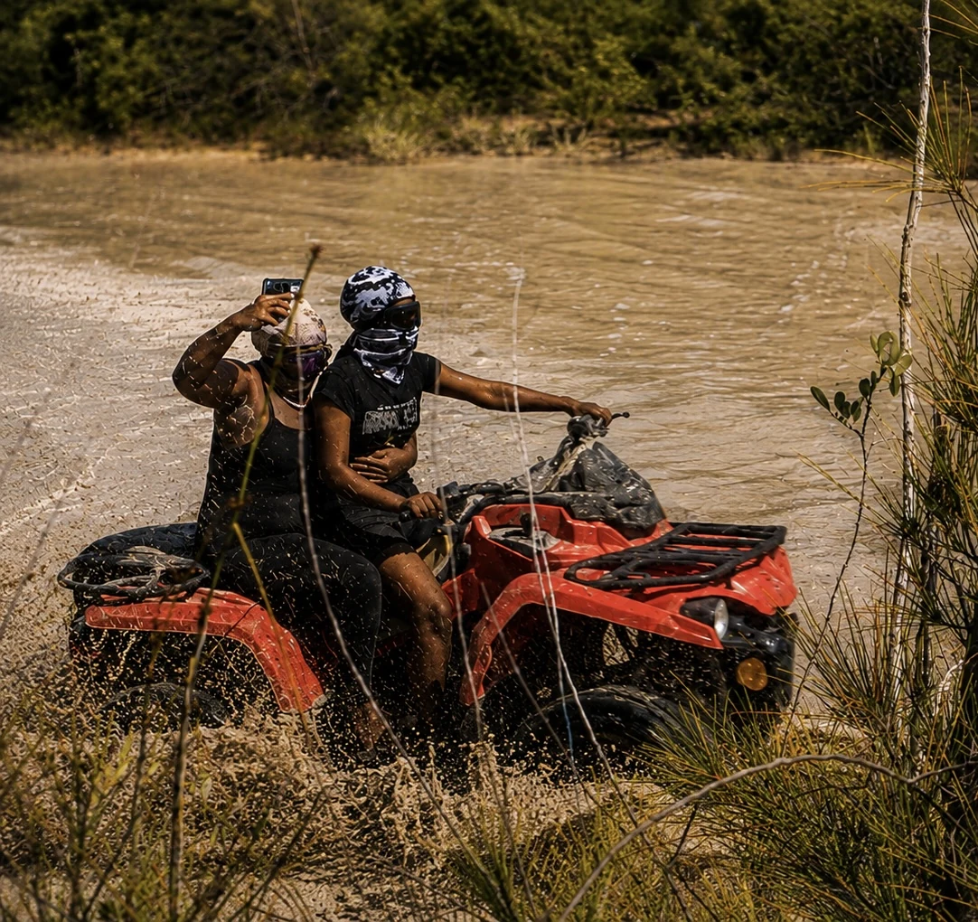 Two riders on a red ATV beside muddy water.
